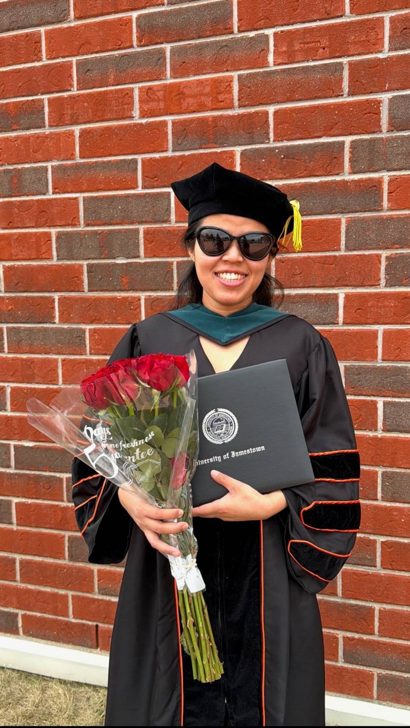 Menuka on graduation day holding her diploma and a bouquet of red roses.